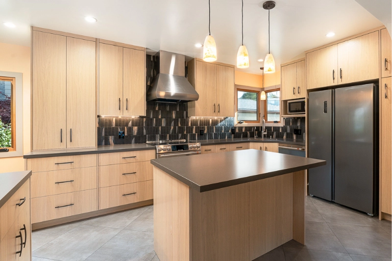 Modern kitchen with light wood cabinets, stainless steel appliances, central island, and black tile backsplash.