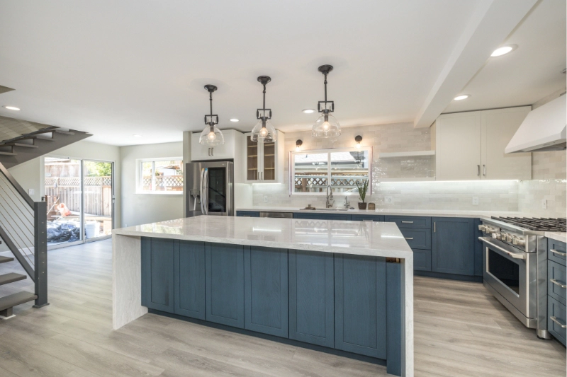 A modern kitchen with a large island featuring blue cabinets, three pendant lights, stainless steel appliances, and light wood flooring.