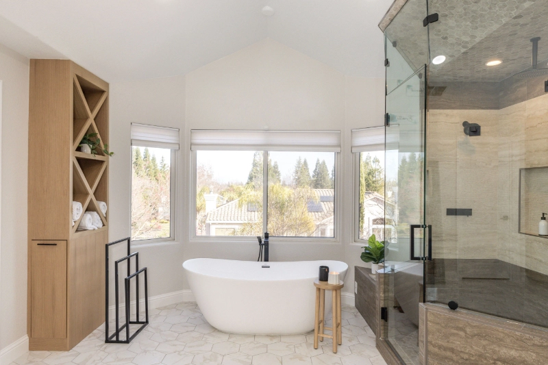 Modern bathroom featuring a freestanding white bathtub, a glass-enclosed shower, a wooden stool, large windows, and a storage unit with shelves.