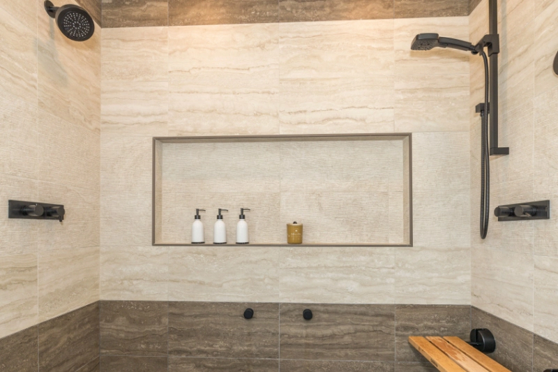 A modern, tiled shower with built-in shelf holding three dispensers and a jar, featuring a rainfall showerhead and a handheld showerhead, with a small wooden bench.