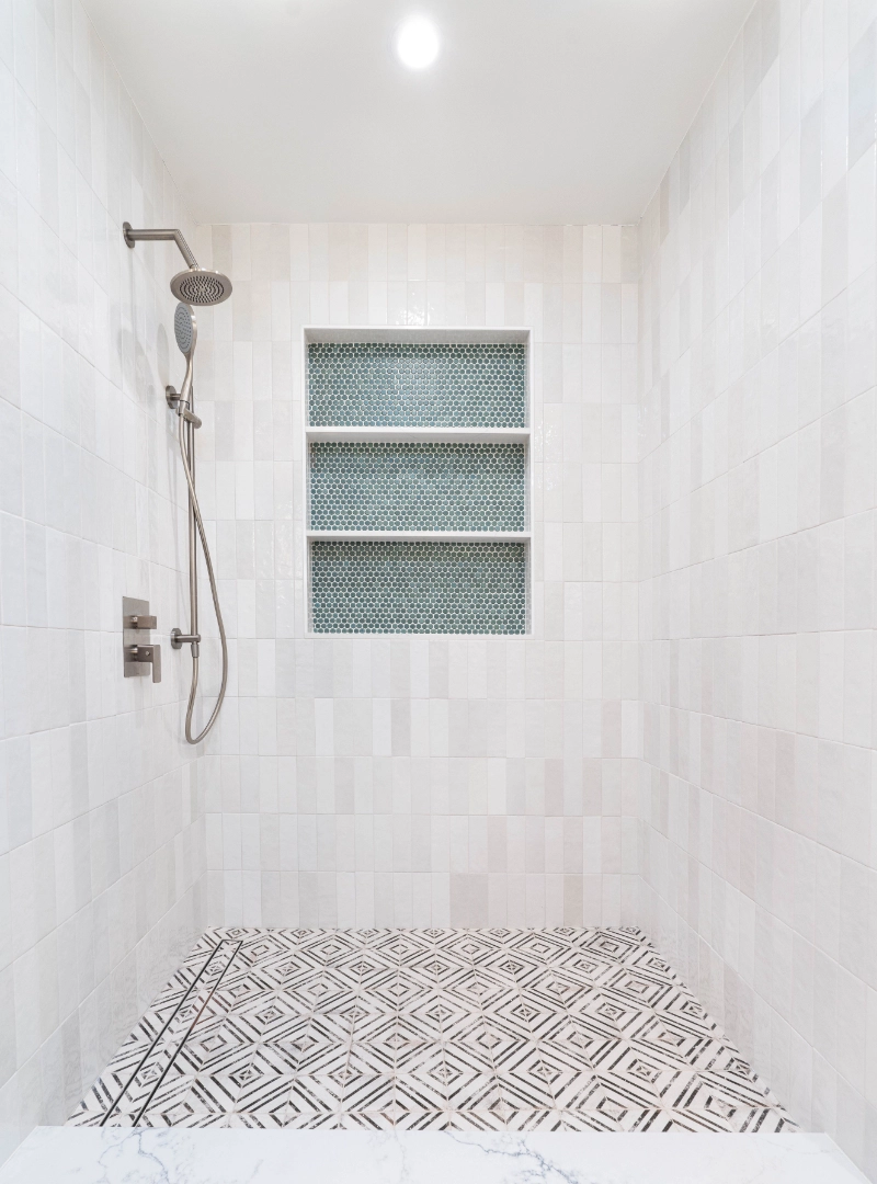 A luxury home remodel featuring a modern shower with white tiled walls, a patterned black and white tiled floor, and a silver-colored showerhead and control panel.