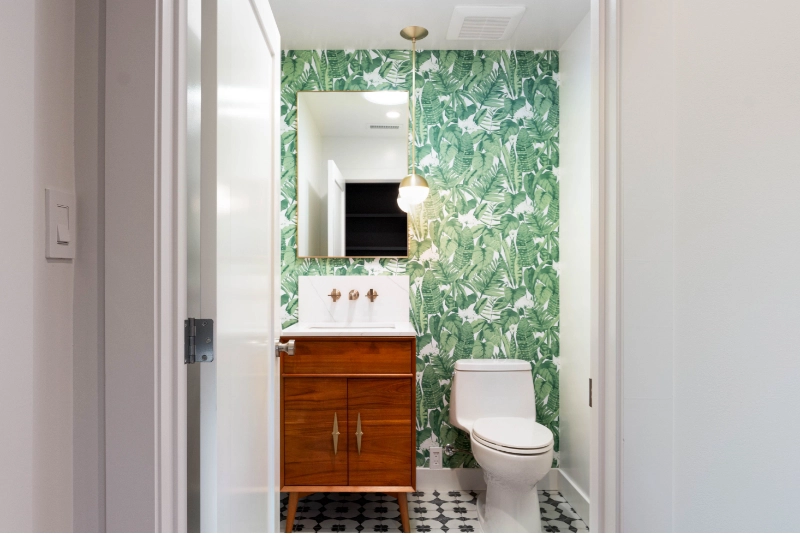 Small bathroom with tropical leaf-patterned wallpaper, white sink with wooden base cabinet, wall-mounted mirror, pendant light, white toilet, and black and white hexagonal floor tiles designed by a luxury home remodel contractor.