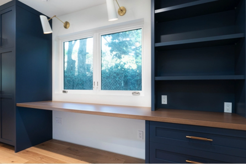 A luxury home remodel featuring a home office with blue cabinetry, a wooden desktop, shelves, and a window overlooking trees.