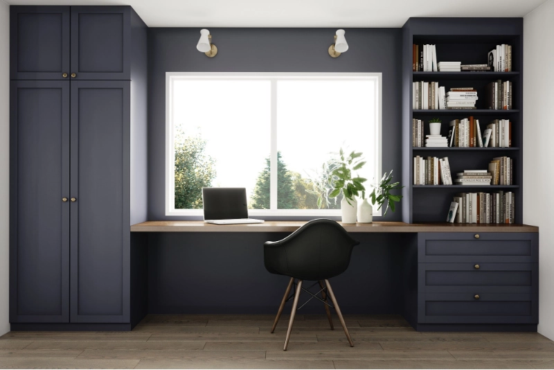 A home office with built-in dark blue cabinets and bookshelves, a wooden desk, a black chair, and a window with outdoor views.
