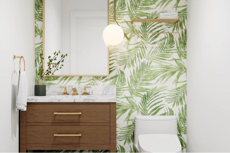A luxurious home remodel features a bathroom with green leaf-patterned wallpaper, a wooden vanity topped with marble, a circular light fixture, gold accents throughout, and a pristine white toilet.