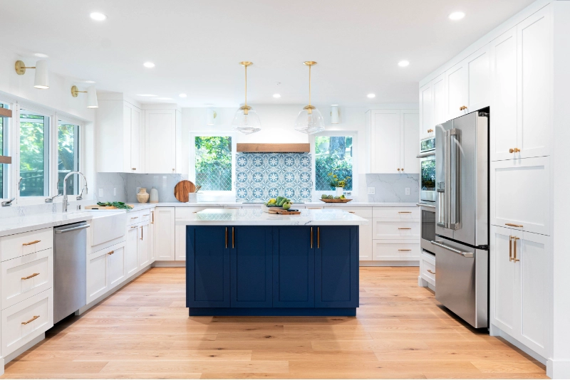 A modern kitchen with white cabinets, a blue island, stainless steel appliances, a light wood floor, and a blue patterned backsplash.