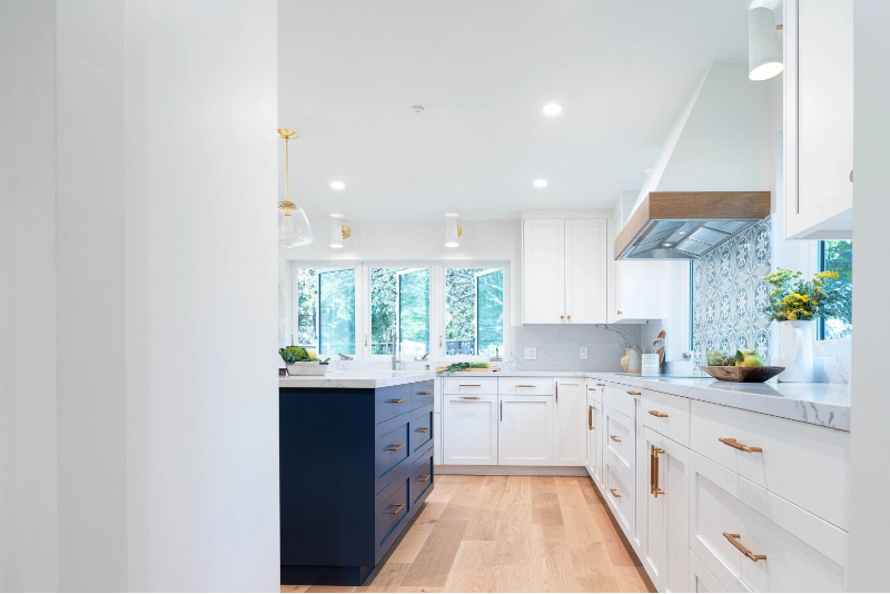 A modern kitchen with white cabinetry, marble countertops, wooden floors, a navy blue island, hanging pendant lights, and large windows providing natural light.