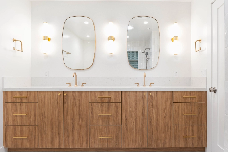 A modern bathroom with two wooden vanities, double sinks, oval mirrors, and gold fixtures.