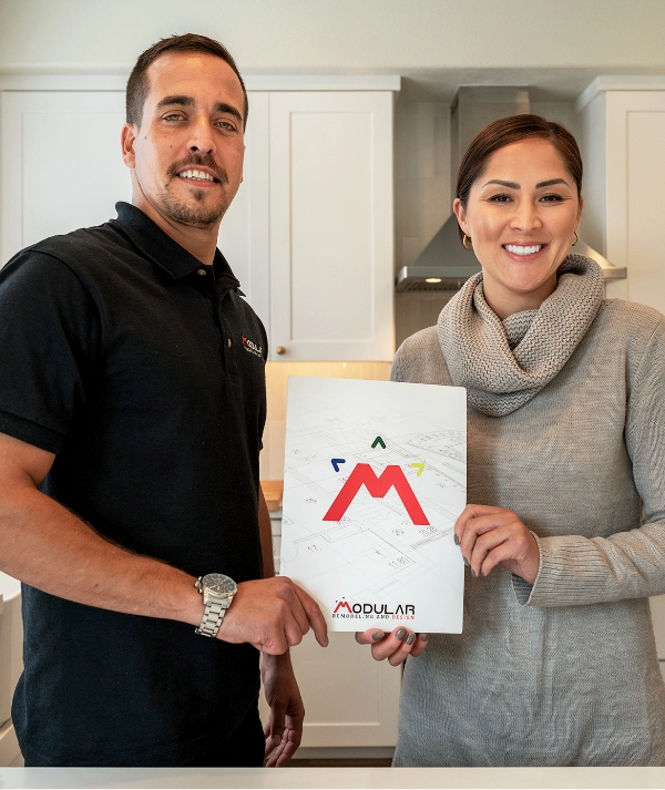A man and woman stand in a kitchen, smiling at the camera and holding a brochure with a red "M" logo and the word "Modular."