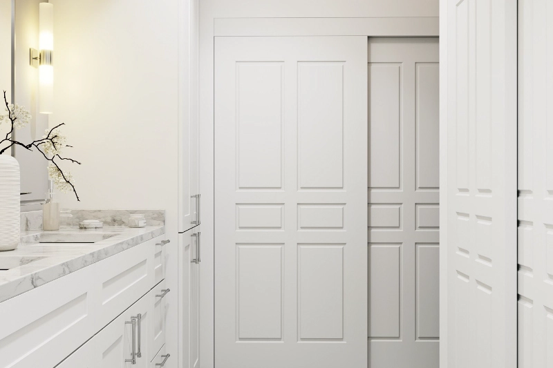 A Regency bathroom remodel features a modern white space with sleek sliding doors, a marble countertop, a sink, cabinets, and a wall-mounted light. The elegant touch is completed with a decorative vase filled with branches.
