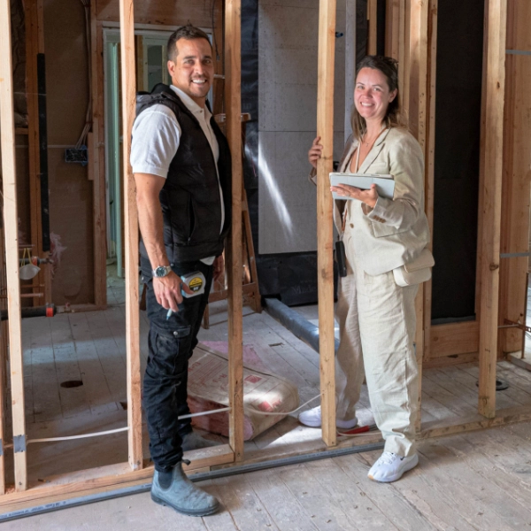 Two people stand inside a house under construction, with exposed wooden framing.
