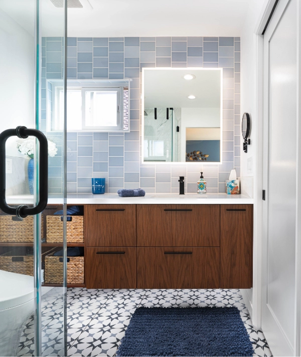 A luxury home remodel showcases a modern bathroom with a wooden vanity and wicker baskets, a lighted mirror, blue and white tiled walls, a glass shower door, and patterned floor tiles.