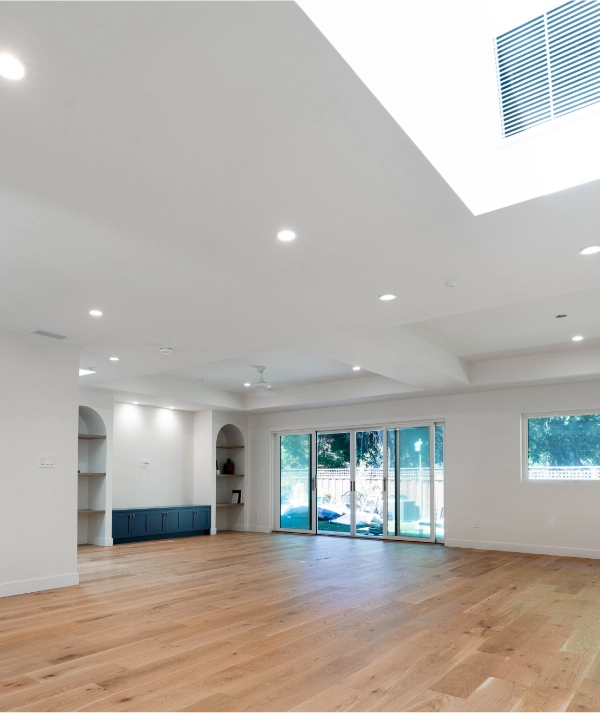 A spacious, empty room with wooden floors, white walls, recessed lighting, a skylight, built-in shelves, and large sliding glass doors leading to an outdoor area.