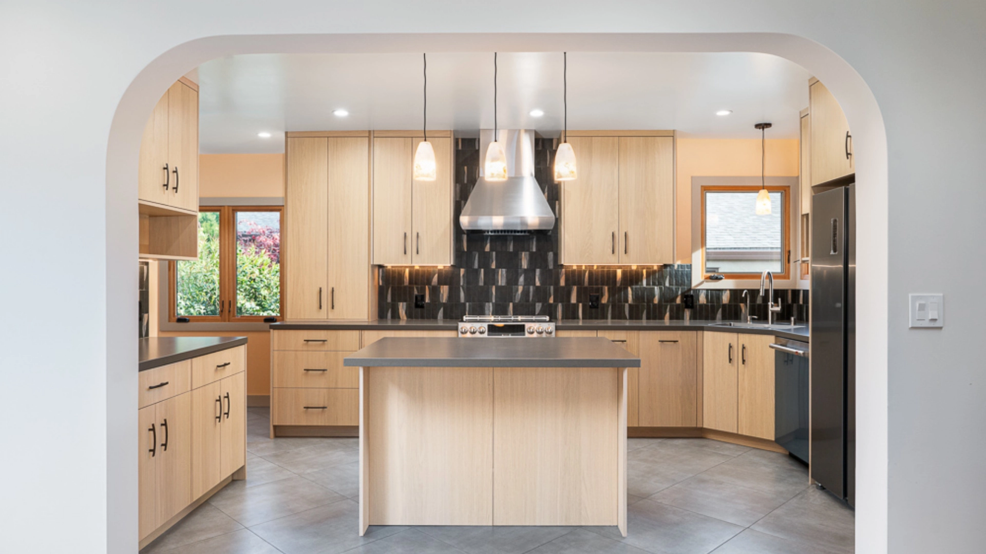 Modern kitchen with light wooden cabinets, dark countertops, a large island, stainless steel appliances, pendant lights, and a black tile backsplash.