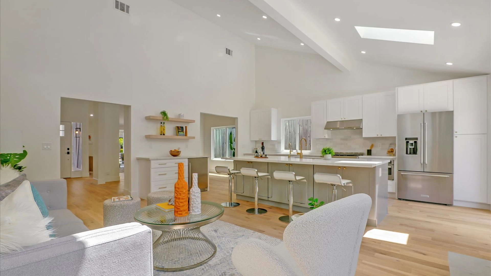 Bright modern kitchen and living area with white cabinets, stainless steel appliances, barstools at the island, and light-colored wooden flooring. An open layout with high ceilings and a skylight.