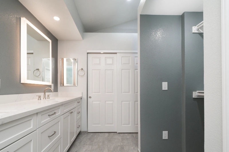Modern Regency bathroom remodel with a gray and white color scheme, featuring a large mirror with built-in lights, double sinks, white cabinets, and sleek white sliding closet doors.